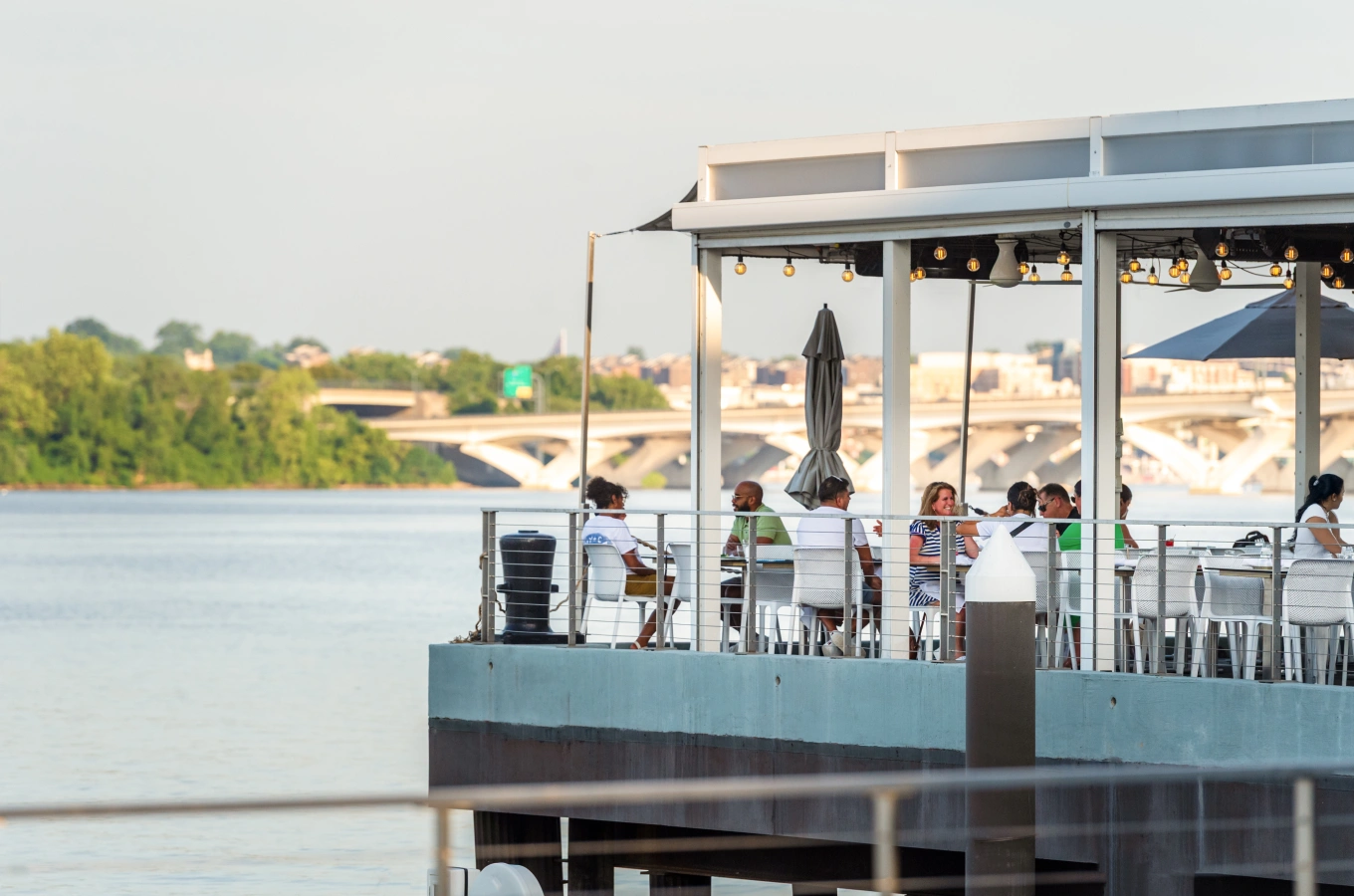 Outdoor waterfront restaurant with people dining under covered patio beside scenic river view
