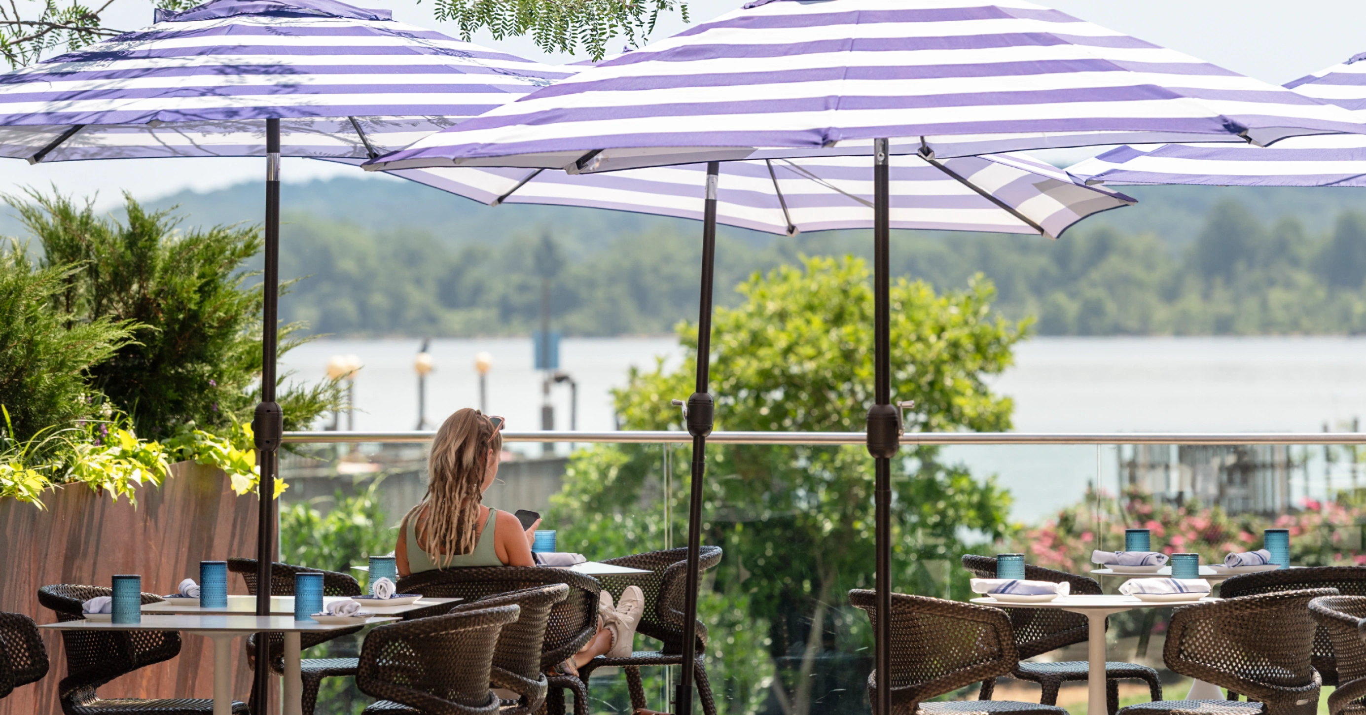 Woman sitting alone at outdoor café with striped umbrellas overlooking lush waterfront view