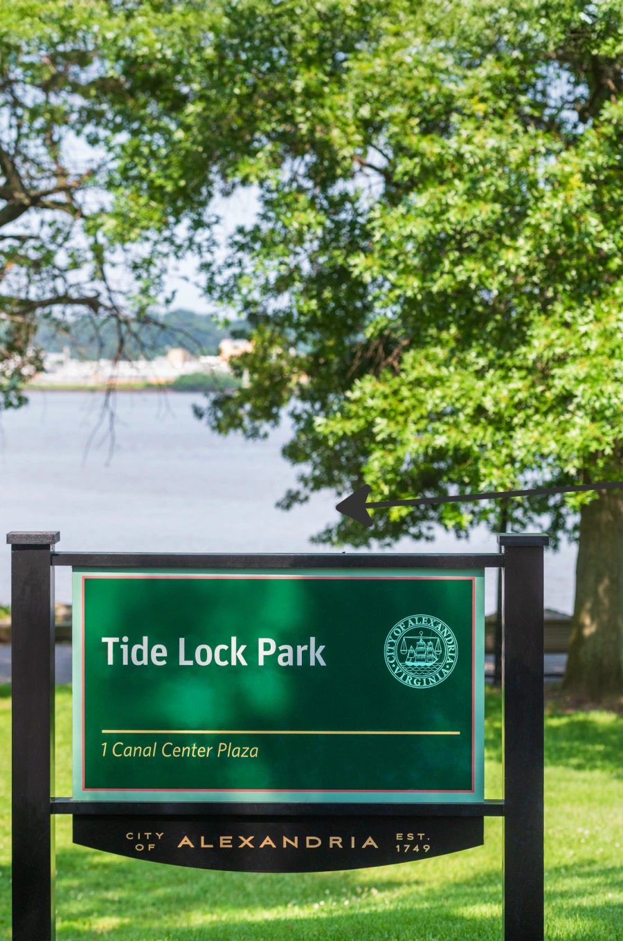 Green park sign for Tide Lock Park with river and leafy trees visible in the scenic background