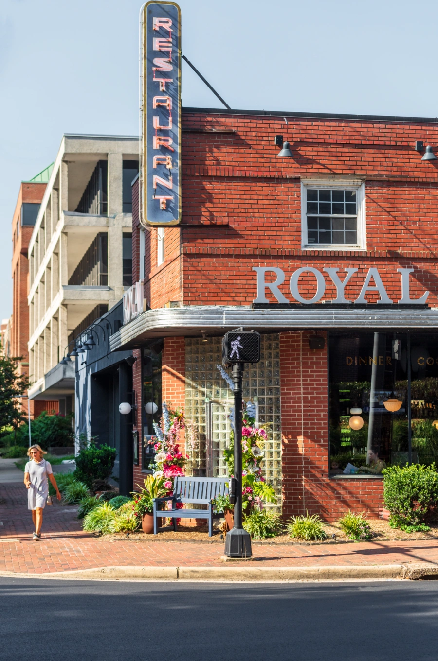 Bright red brick corner building labeled Royal with neon restaurant sign and lush sidewalk plants